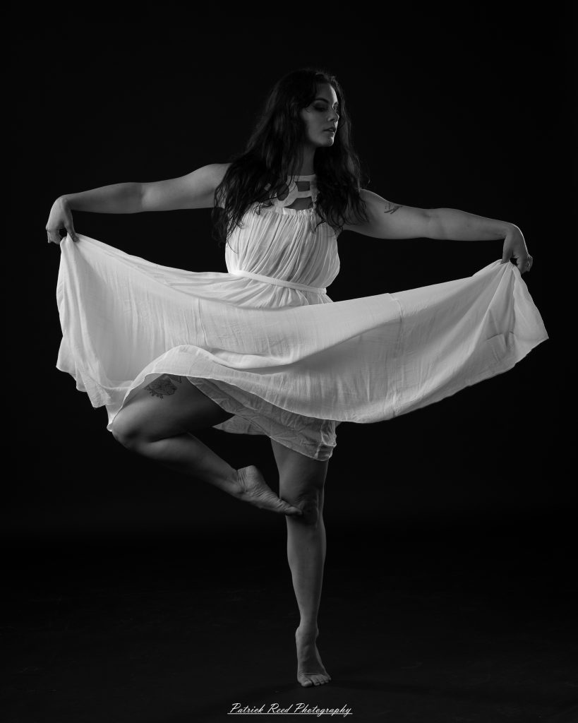 Black and white fine art dance portrait of a dancer in a flowing dress, captured in motion with expressive movement and soft studio lighting against a dark background.