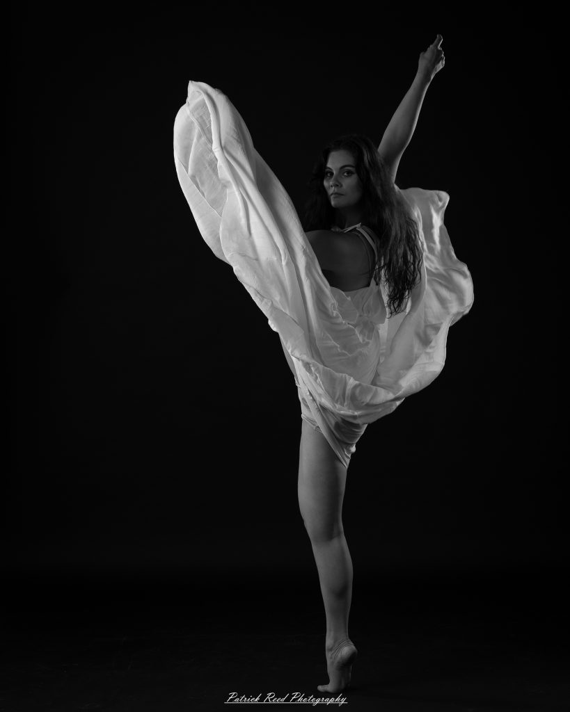 Black and white fine art dance portrait of a dancer in a flowing dress, captured in motion with expressive movement and soft studio lighting against a dark background.