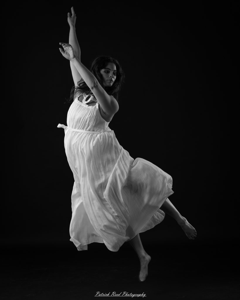 Black and white fine art dance portrait of a dancer in a flowing dress, captured in motion with expressive movement and soft studio lighting against a dark background.