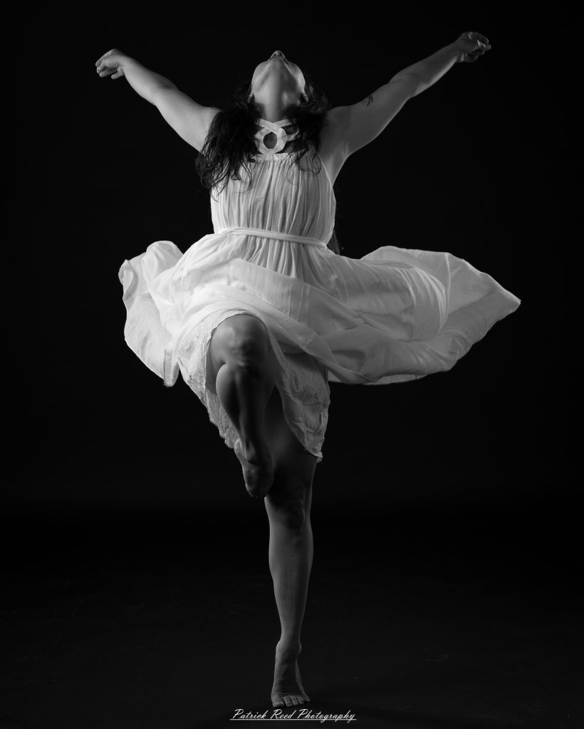 Black and white fine art dance portrait of a dancer in a flowing dress, captured in motion with expressive movement and soft studio lighting against a dark background.