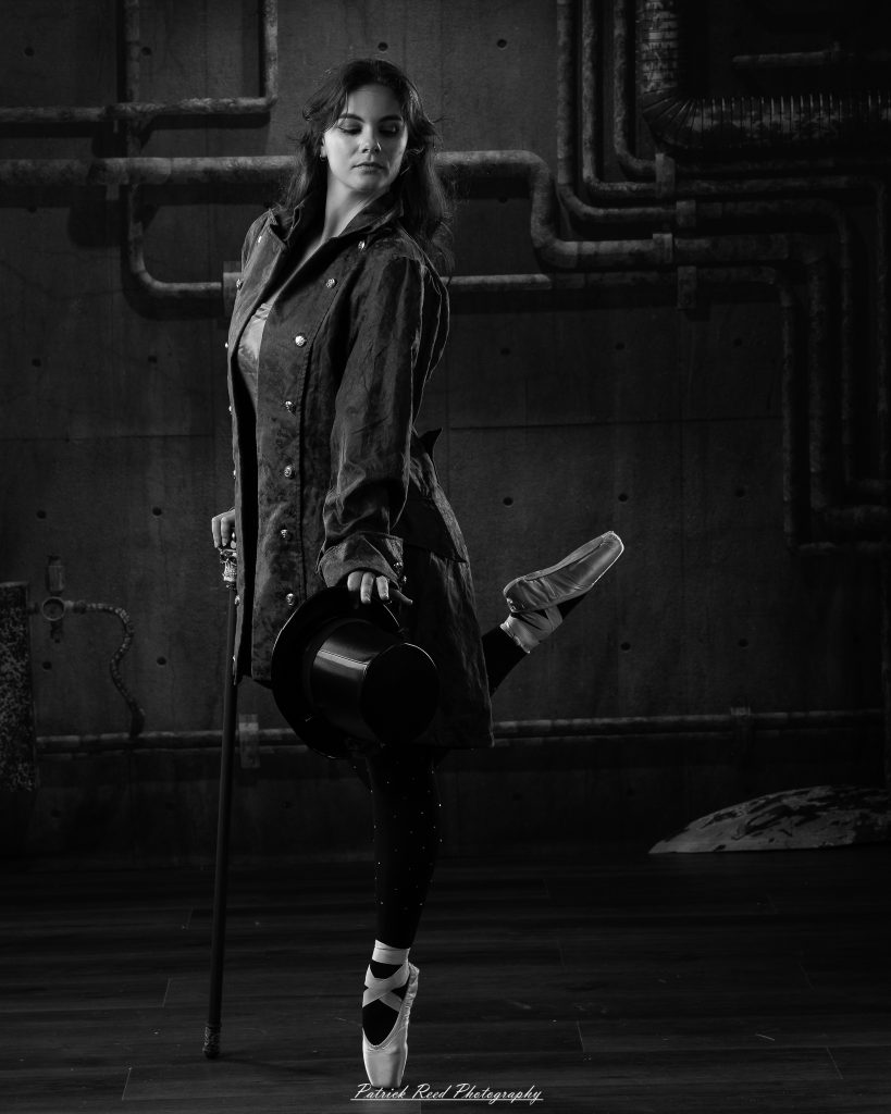 Black and white portrait of a steampunk-inspired dancer wearing a coat, hat, and holding a cane, performing expressive movements in an industrial studio setting with pipes and dramatic lighting.
