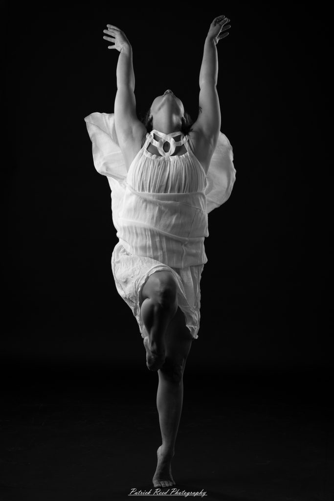Black and white fine art dance portrait of a dancer in a flowing dress, captured in motion with expressive movement and soft studio lighting against a dark background.