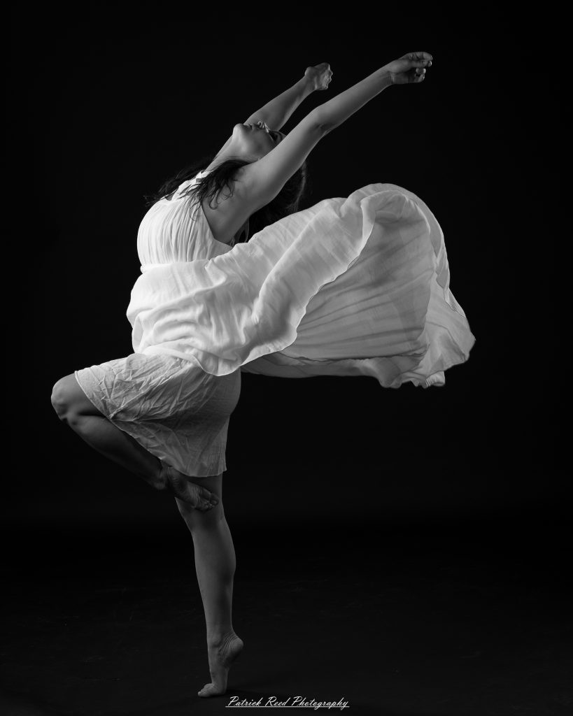 Black and white fine art dance portrait of a dancer in a flowing dress, captured in motion with expressive movement and soft studio lighting against a dark background.