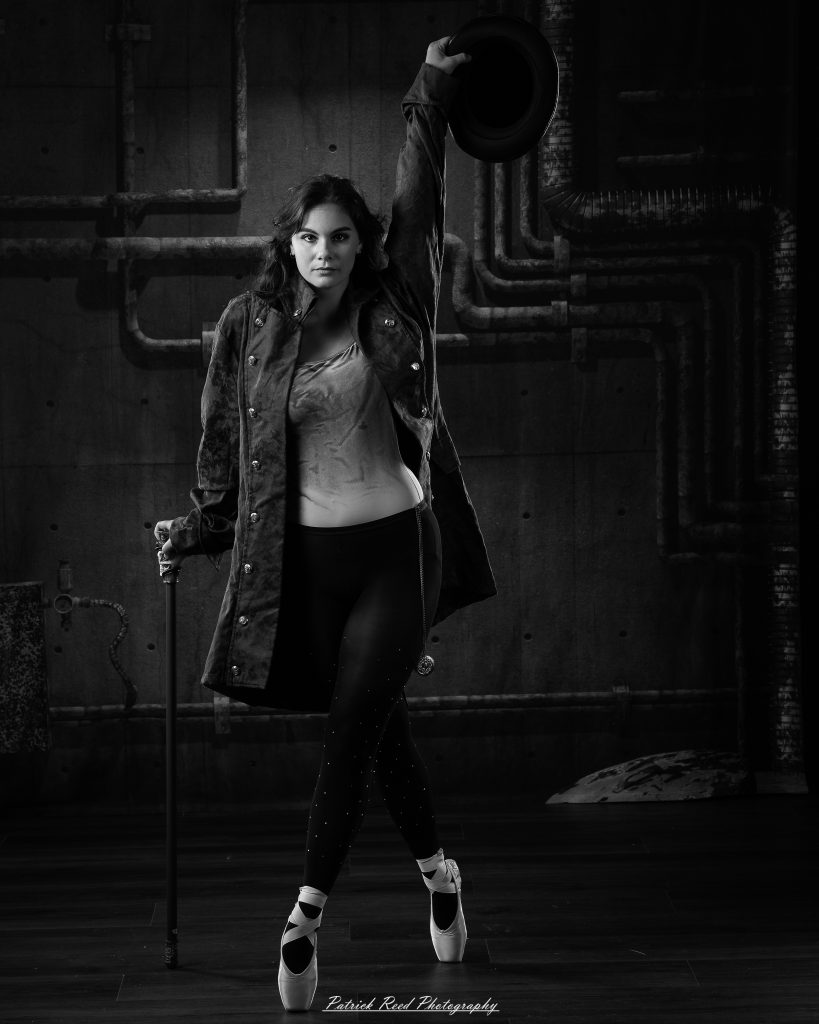 Black and white portrait of a steampunk-inspired dancer wearing a coat, hat, and holding a cane, performing expressive movements in an industrial studio setting with pipes and dramatic lighting.
