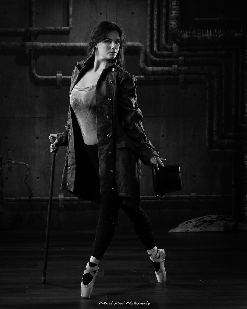 Black and white portrait of a steampunk-inspired dancer wearing a coat, hat, and holding a cane, performing expressive movements in an industrial studio setting with pipes and dramatic lighting.