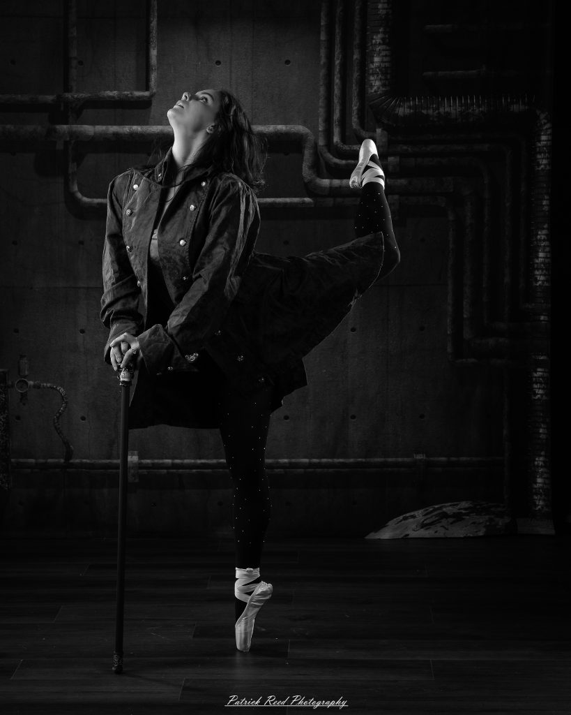 Black and white portrait of a steampunk-inspired dancer wearing a coat, hat, and holding a cane, performing expressive movements in an industrial studio setting with pipes and dramatic lighting.