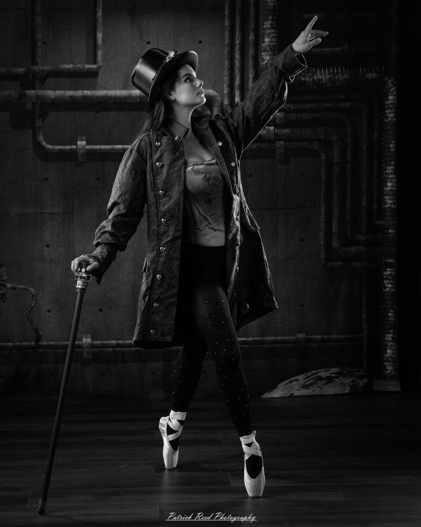 Black and white portrait of a steampunk-inspired dancer wearing a coat, hat, and holding a cane, performing expressive movements in an industrial studio setting with pipes and dramatic lighting.