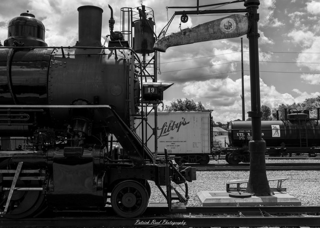 Black-and-white documentary photographs from the Steam Railroading Institute in Ohio, featuring historic steam locomotives, vintage passenger railcars, railroad machinery, restoration workshops, and industrial details preserving America's railroad heritage.