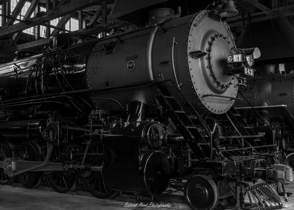Black-and-white documentary photographs from the Steam Railroading Institute in Ohio, featuring historic steam locomotives, vintage passenger railcars, railroad machinery, restoration workshops, and industrial details preserving America's railroad heritage.