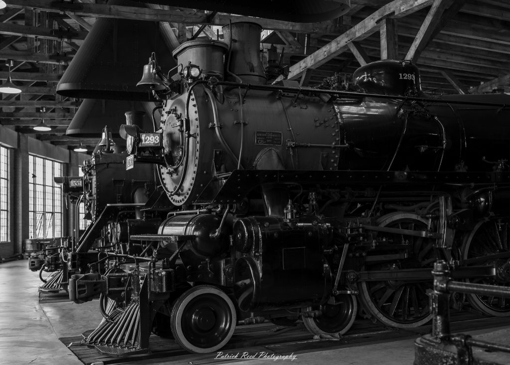 Black-and-white documentary photographs from the Steam Railroading Institute in Ohio, featuring historic steam locomotives, vintage passenger railcars, railroad machinery, restoration workshops, and industrial details preserving America's railroad heritage.