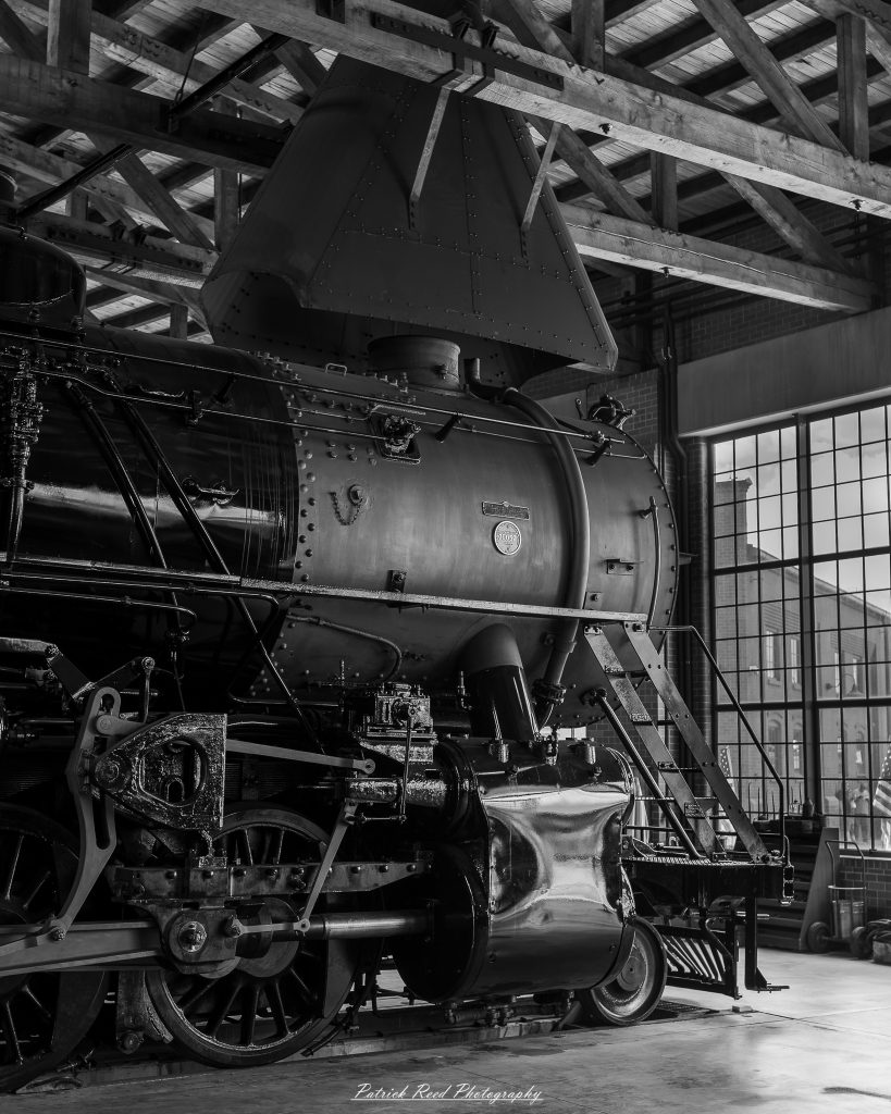 Black-and-white documentary photographs from the Steam Railroading Institute in Ohio, featuring historic steam locomotives, vintage passenger railcars, railroad machinery, restoration workshops, and industrial details preserving America's railroad heritage.