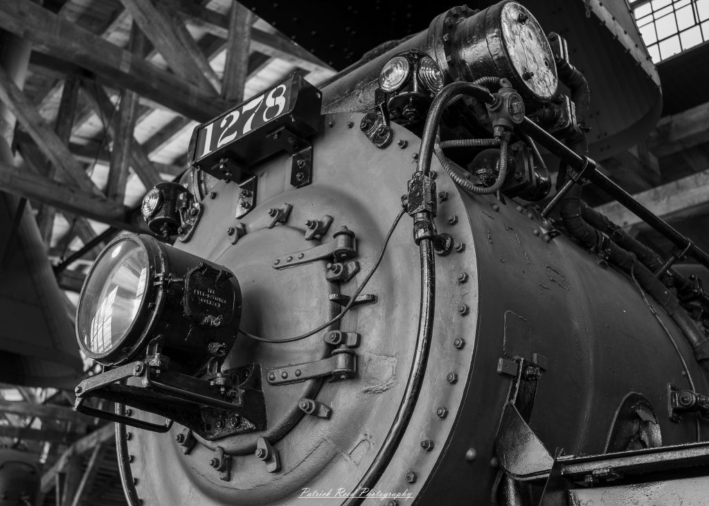 Black-and-white documentary photographs from the Steam Railroading Institute in Ohio, featuring historic steam locomotives, vintage passenger railcars, railroad machinery, restoration workshops, and industrial details preserving America's railroad heritage.