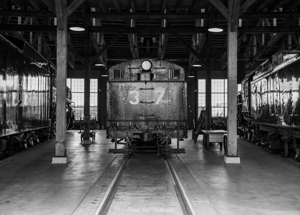 Black-and-white documentary photographs from the Steam Railroading Institute in Ohio, featuring historic steam locomotives, vintage passenger railcars, railroad machinery, restoration workshops, and industrial details preserving America's railroad heritage.
