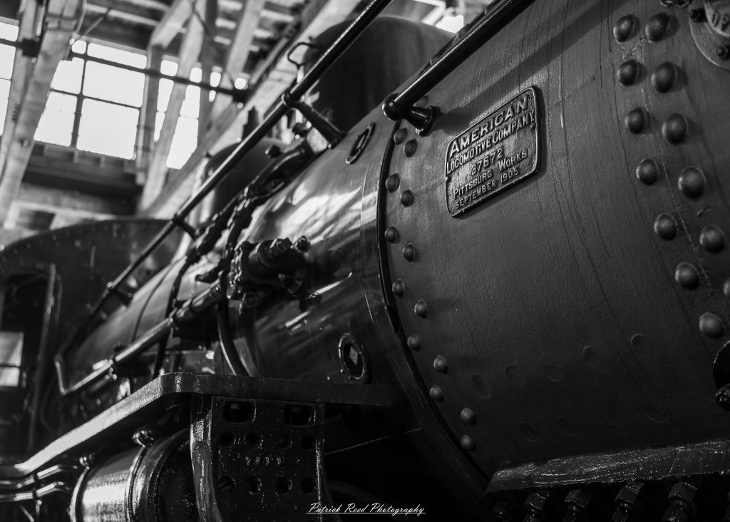 Black-and-white documentary photographs from the Steam Railroading Institute in Ohio, featuring historic steam locomotives, vintage passenger railcars, railroad machinery, restoration workshops, and industrial details preserving America's railroad heritage.