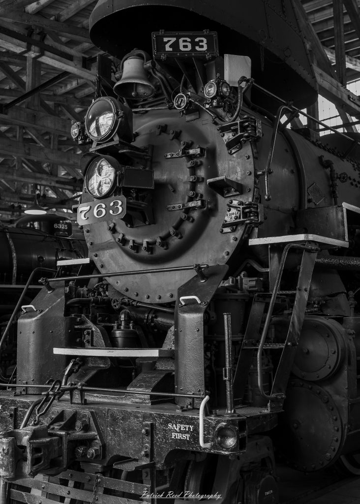 Black-and-white documentary photographs from the Steam Railroading Institute in Ohio, featuring historic steam locomotives, vintage passenger railcars, railroad machinery, restoration workshops, and industrial details preserving America's railroad heritage.