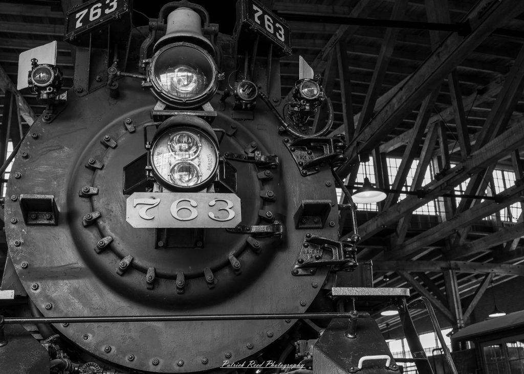Black-and-white documentary photographs from the Steam Railroading Institute in Ohio, featuring historic steam locomotives, vintage passenger railcars, railroad machinery, restoration workshops, and industrial details preserving America's railroad heritage.