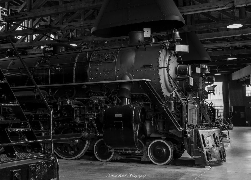 Black-and-white documentary photographs from the Steam Railroading Institute in Ohio, featuring historic steam locomotives, vintage passenger railcars, railroad machinery, restoration workshops, and industrial details preserving America's railroad heritage.