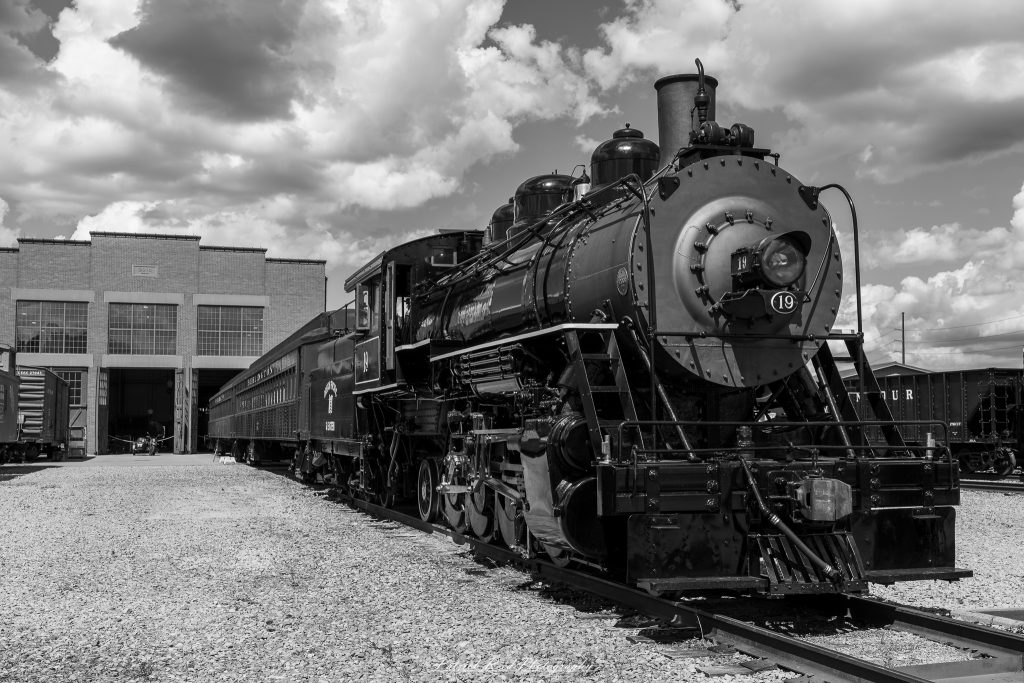 Black-and-white documentary photographs from the Steam Railroading Institute in Ohio, featuring historic steam locomotives, vintage passenger railcars, railroad machinery, restoration workshops, and industrial details preserving America's railroad heritage.