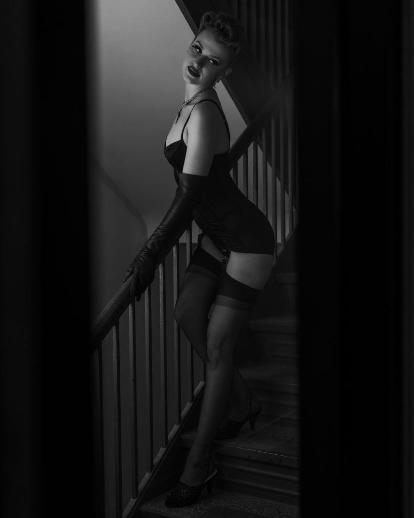 Black-and-white portrait of a woman in lingerie, gloves, and stockings posing on a stair railing.