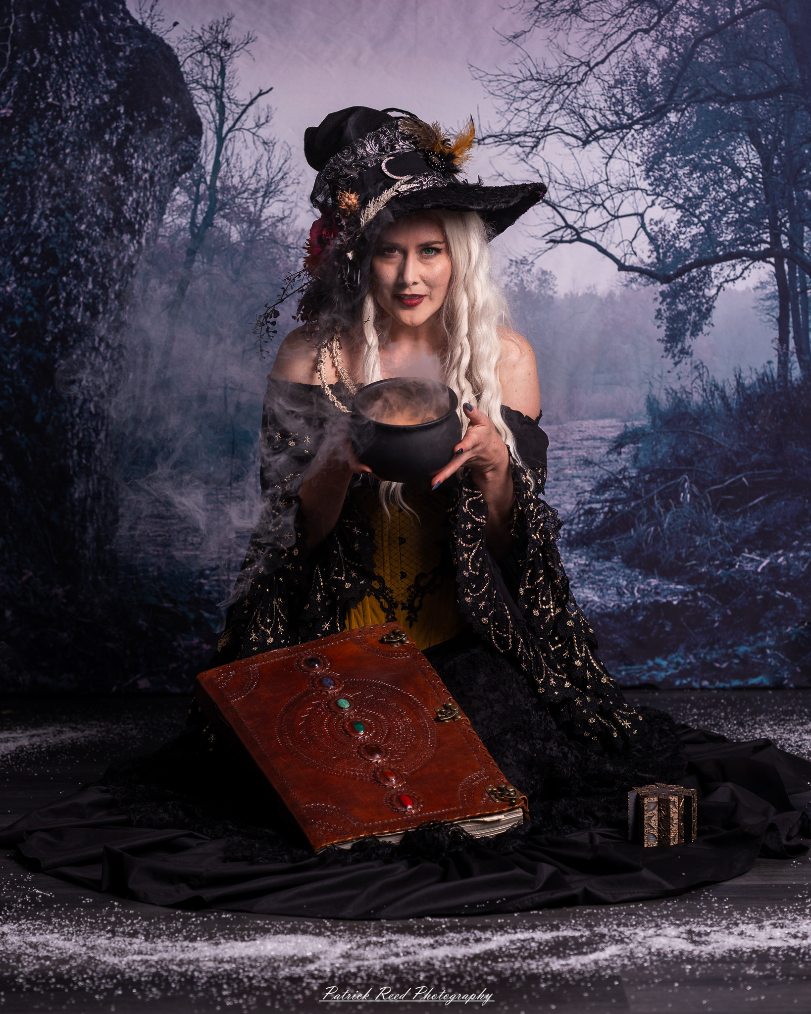 Woman dressed as a witch in a decorated black hat and lace gown, holding a steaming black cauldron above an ornate spellbook, against a misty forest backdrop.