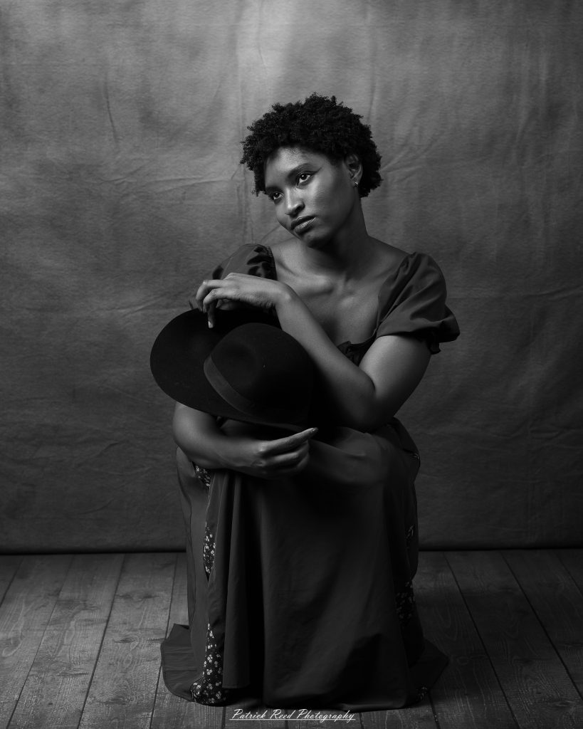 Black-and-white portrait of a woman in a dark off-the-shoulder dress, seated against a draped backdrop.