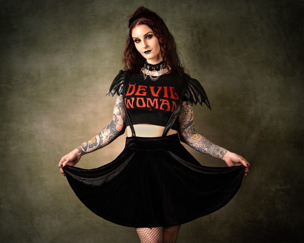Tattooed woman with red hair, black choker and leather jacket, holding skirt in studio portrait.