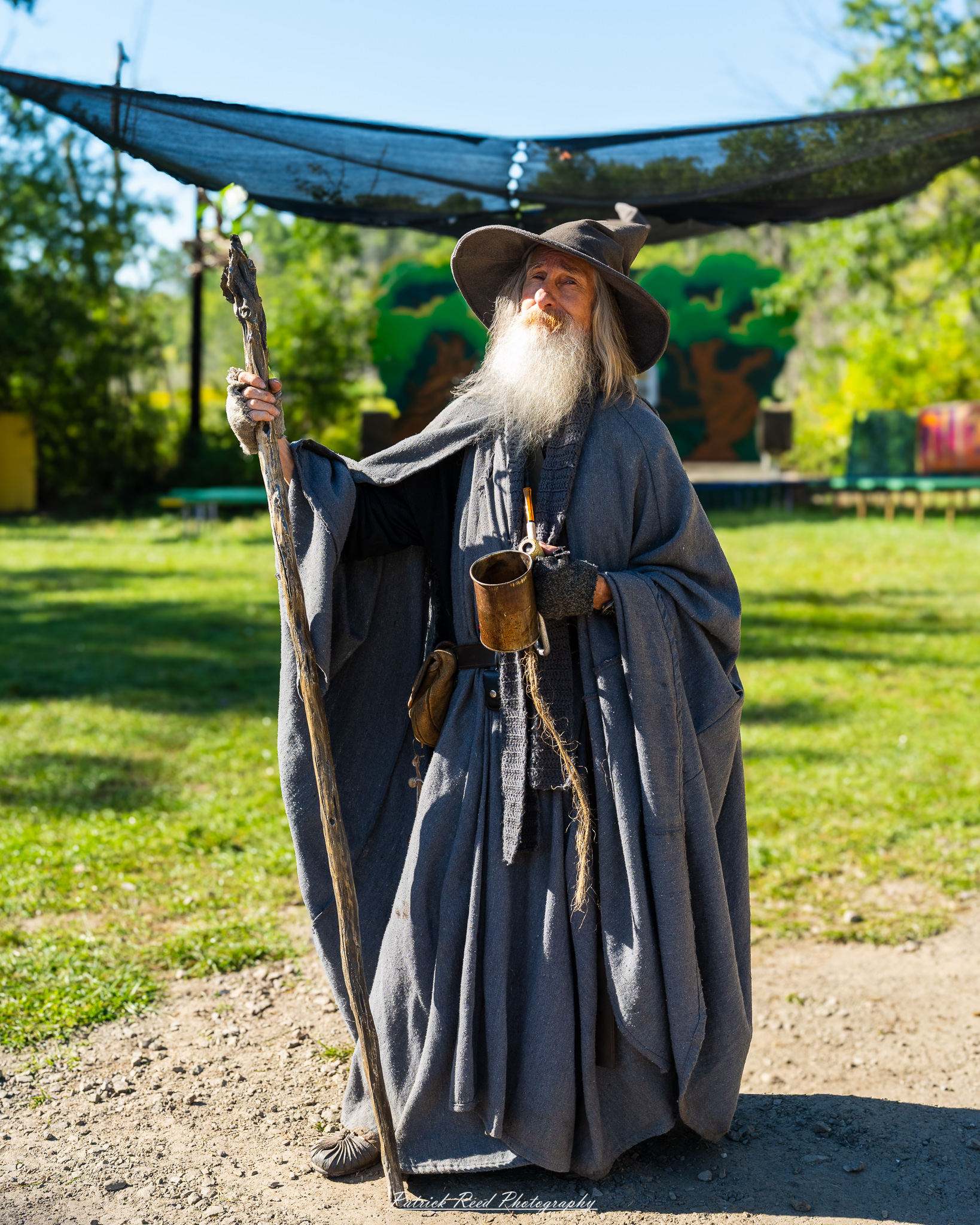 Historical reenactor in dark robe and hat holding a spear at a Renaissance fair camp.