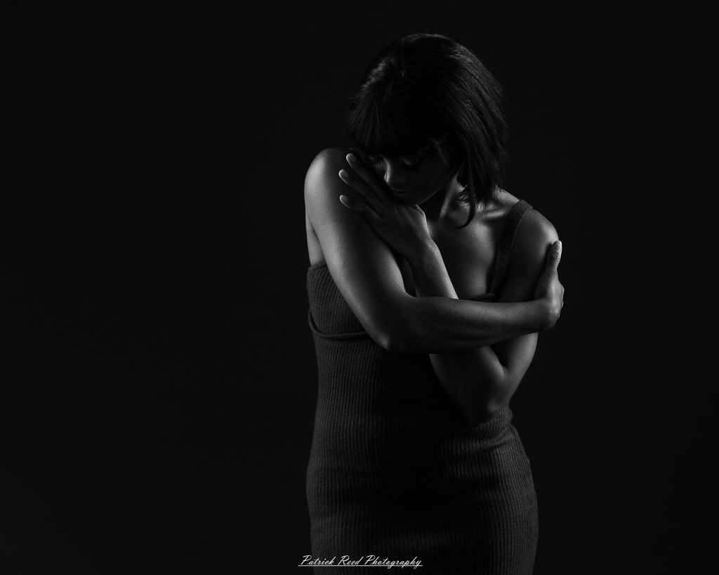 Black-and-white portrait of a woman looking down with her arms crossed over her chest, lit against a dark background.