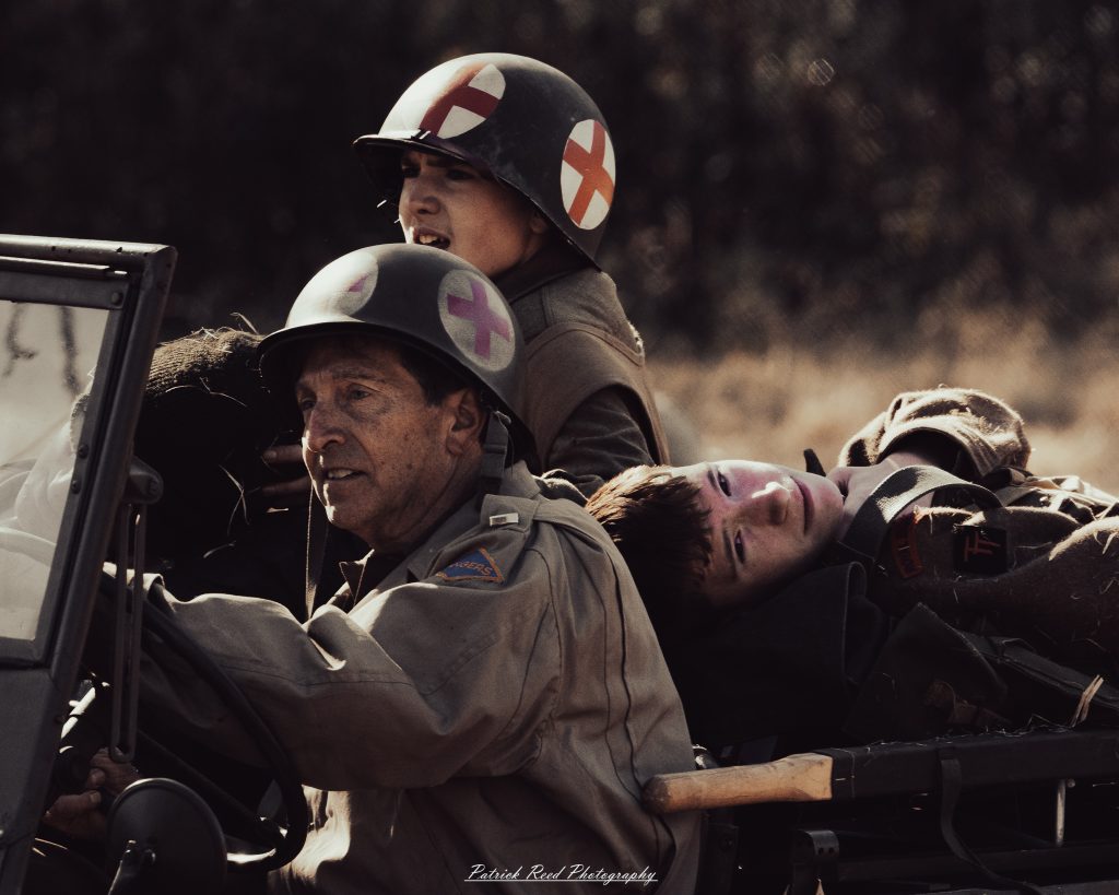 Two paramedics in an ambulance wearing helmets with red crosses during a historical reenactment transport a wounded soldier