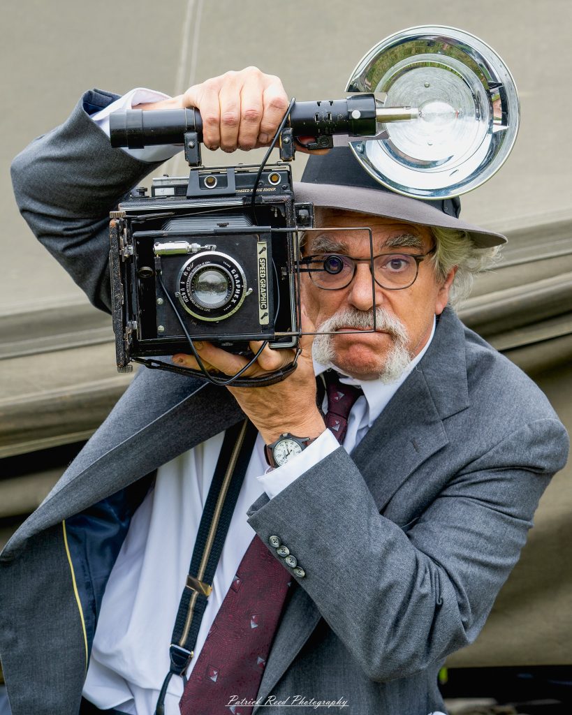 Man in a suit and fedora aiming a vintage Speed Graphic camera with a large flash reflector.