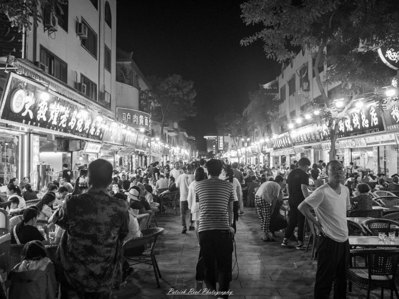 Black-and-white photograph of a crowded Chinese food street at night, with diners seated beneath illuminated restaurant signs on both sides.