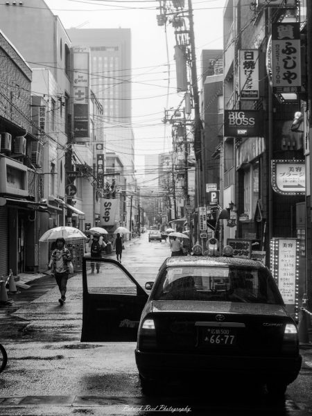 "A black and white street photography scene in Japan captures the timeless essence of urban life. The image features pedestrians walking along a busy street, with a mix of traditional and modern architecture in the background. Neon signs and advertisements, though muted in grayscale, still convey the vibrant energy of the city. The contrast between light and shadow highlights the textures and details of the street, from the patterns on the pavement to the clothing of the passersby. The overall mood is one of quiet observation, focusing on the everyday interactions and movements within the urban landscape."
