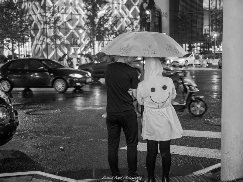 Black-and-white photograph of two people sharing an umbrella beside wet nighttime traffic, one wearing a hooded coat with a large smiley face.