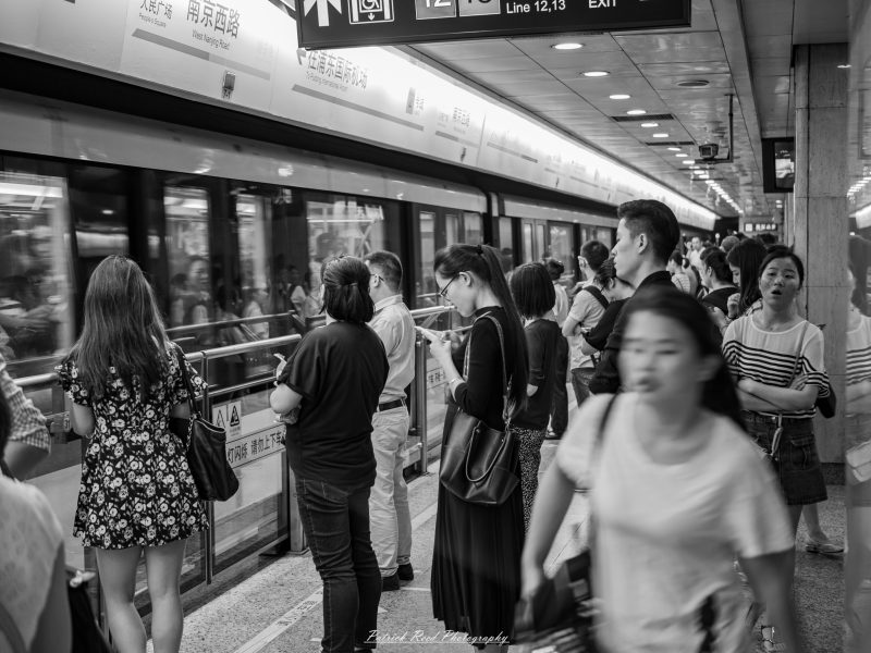 Black-and-white photograph of commuters waiting beside a moving Shanghai Metro train, with a woman looking at her phone near the platform railing.