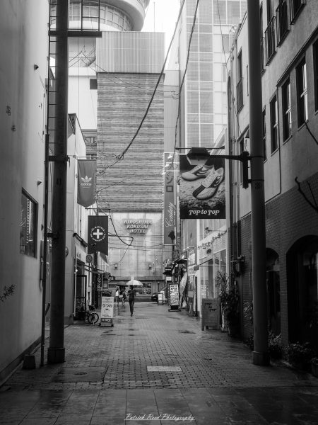 Black-and-white view down a narrow Hiroshima shopping alley, with hanging storefront signs and an umbrella-carrying pedestrian.