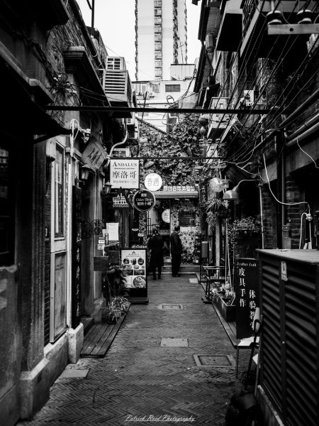 Black-and-white view down a narrow Shanghai shopping lane lined with brick walls, hanging signs, overhead wires and small storefronts.