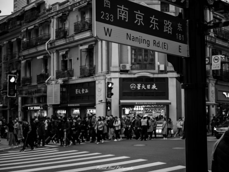 Black-and-white photograph of pedestrians crossing beneath a Nanjing Road East street sign in Shanghai.