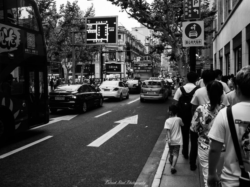 Black-and-white photograph of pedestrians, including an adult holding a child’s hand, walking beside buses and cars on a busy Shanghai street.