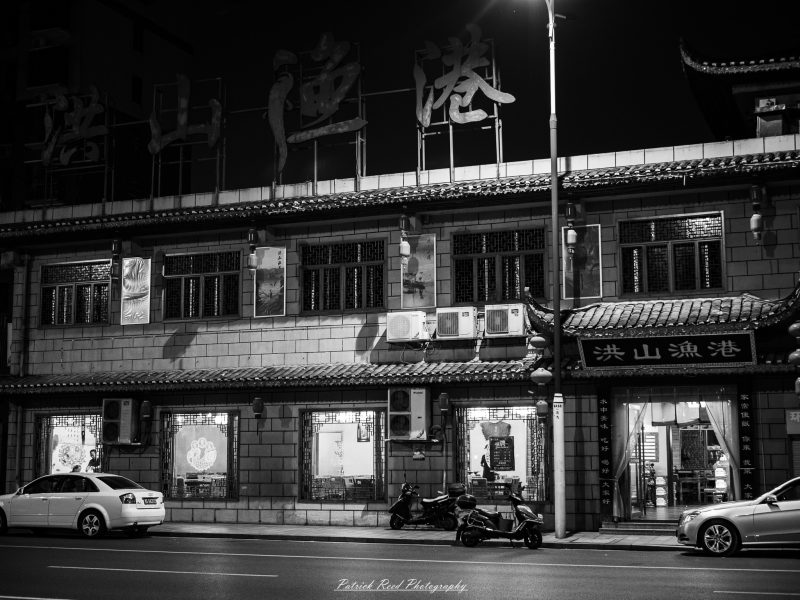 Black-and-white nighttime photograph of a Shanghai restaurant with illuminated windows, large Chinese roof signs and scooters parked outside.