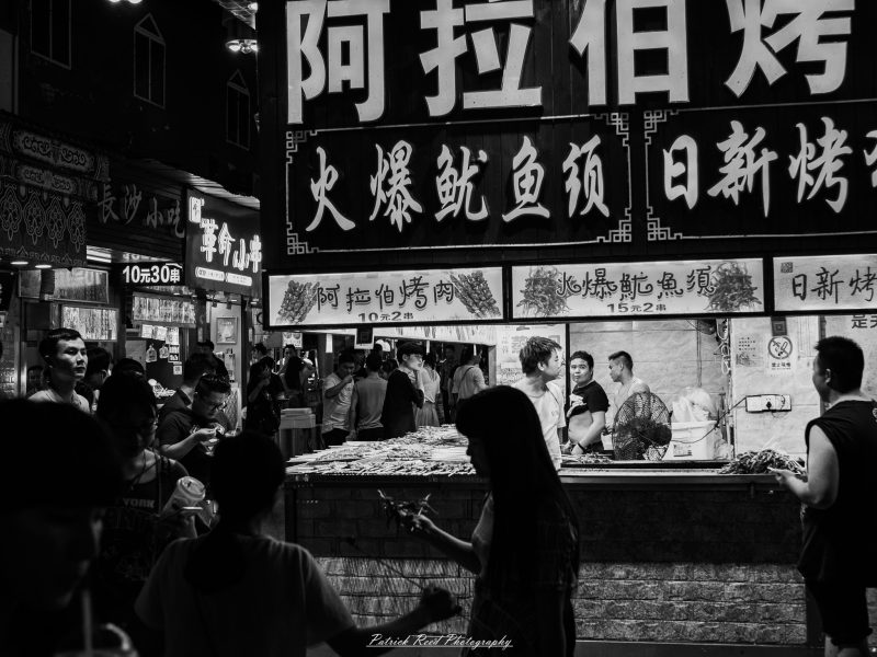 Black-and-white photograph of customers passing a Shanghai food stall beneath large Chinese signs, with vendors working behind rows of skewers.