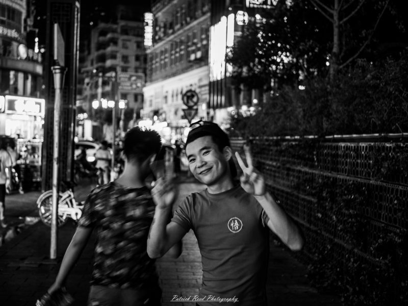 A smiling man flashes two peace signs on a Shanghai sidewalk at night in a black-and-white street photograph.