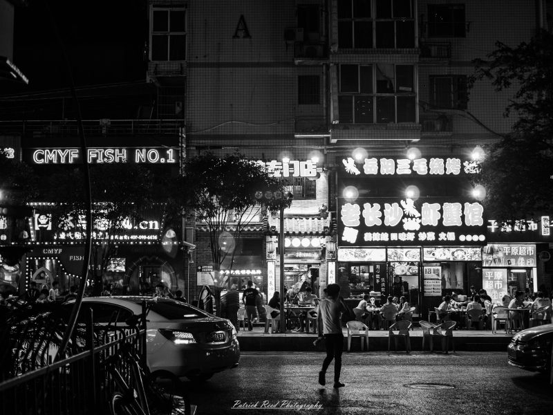Black-and-white nighttime photograph of a pedestrian crossing in front of brightly lit Shanghai restaurants and outdoor diners.