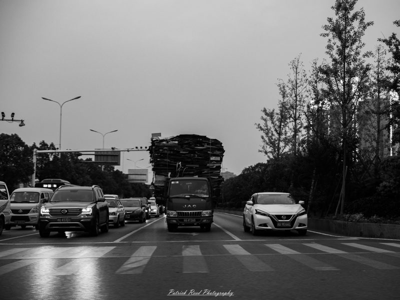 A truck carrying a tall stack of flattened material approaches a Shanghai intersection among cars in a black-and-white photograph.