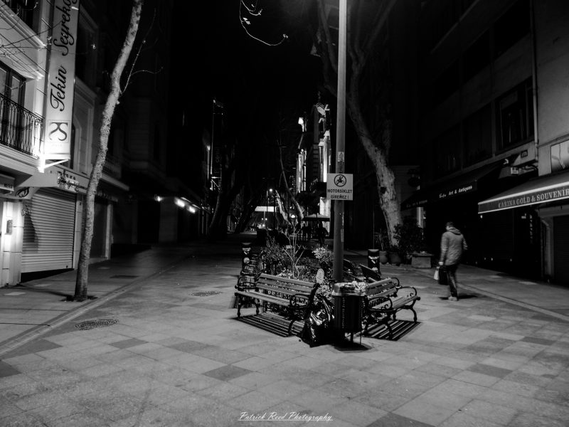 A lone pedestrian passes shuttered shops and empty benches on an Istanbul street at night in a black-and-white photograph.