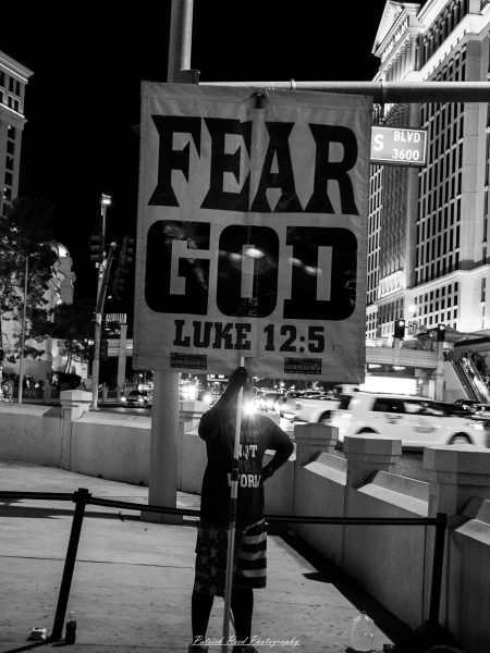 A person holds a large “FEAR GOD — LUKE 12:5” sign beside nighttime traffic on the Las Vegas Strip, photographed in black and white.