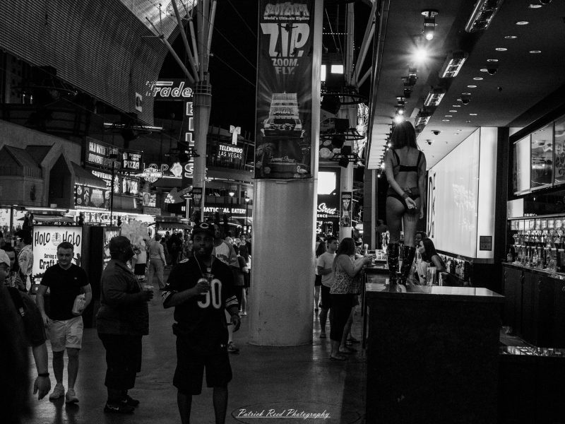 Pedestrians pass an outdoor bar where a dancer stands on a platform beneath casino lights in a black-and-white Las Vegas street photograph.