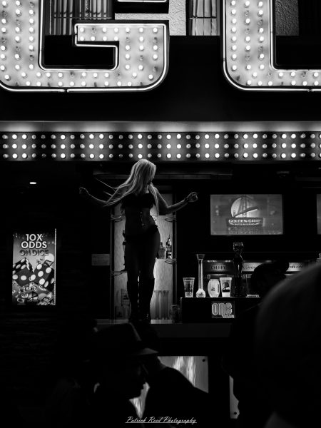 A dancer stands beneath bright casino marquee bulbs, with passing figures silhouetted below, in a black-and-white Las Vegas photograph.