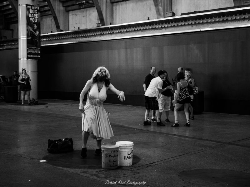 Black-and-white photograph of a bearded street performer in a light halter dress and wig beside tip buckets in Las Vegas.