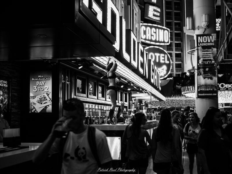 Black-and-white photograph of a dancer on an outdoor platform beneath Golden Gate casino signs as pedestrians pass in Las Vegas.