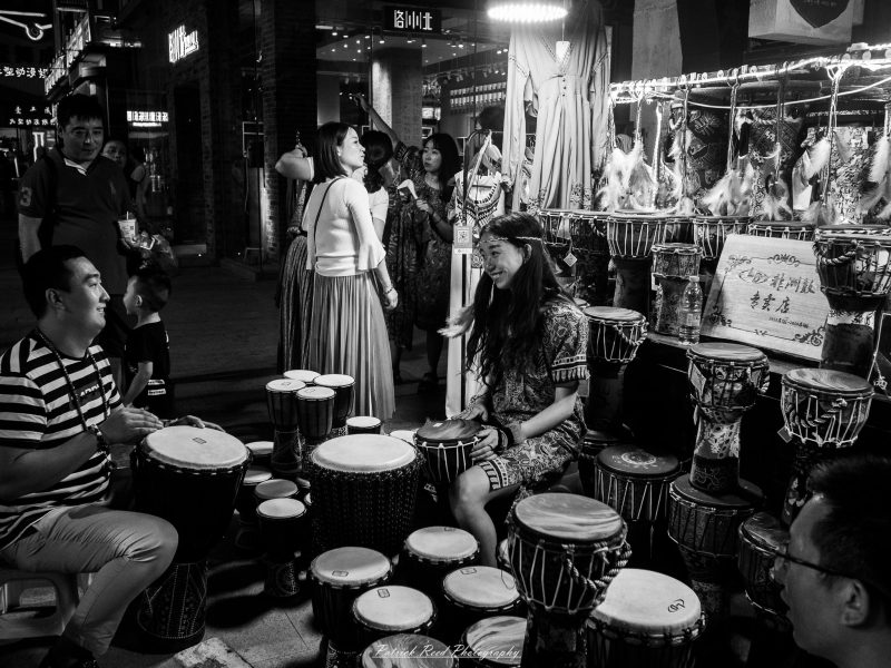Black-and-white photograph of a smiling woman and man playing hand drums amid rows of drums at a Chinese market stall.